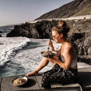 Laura Schiller, von anotherlivingbeing, sitzend auf einer Mauer vor einer Klippe. Im Hintergrund sieht man die Wellen und die Vulkane von La Palma.