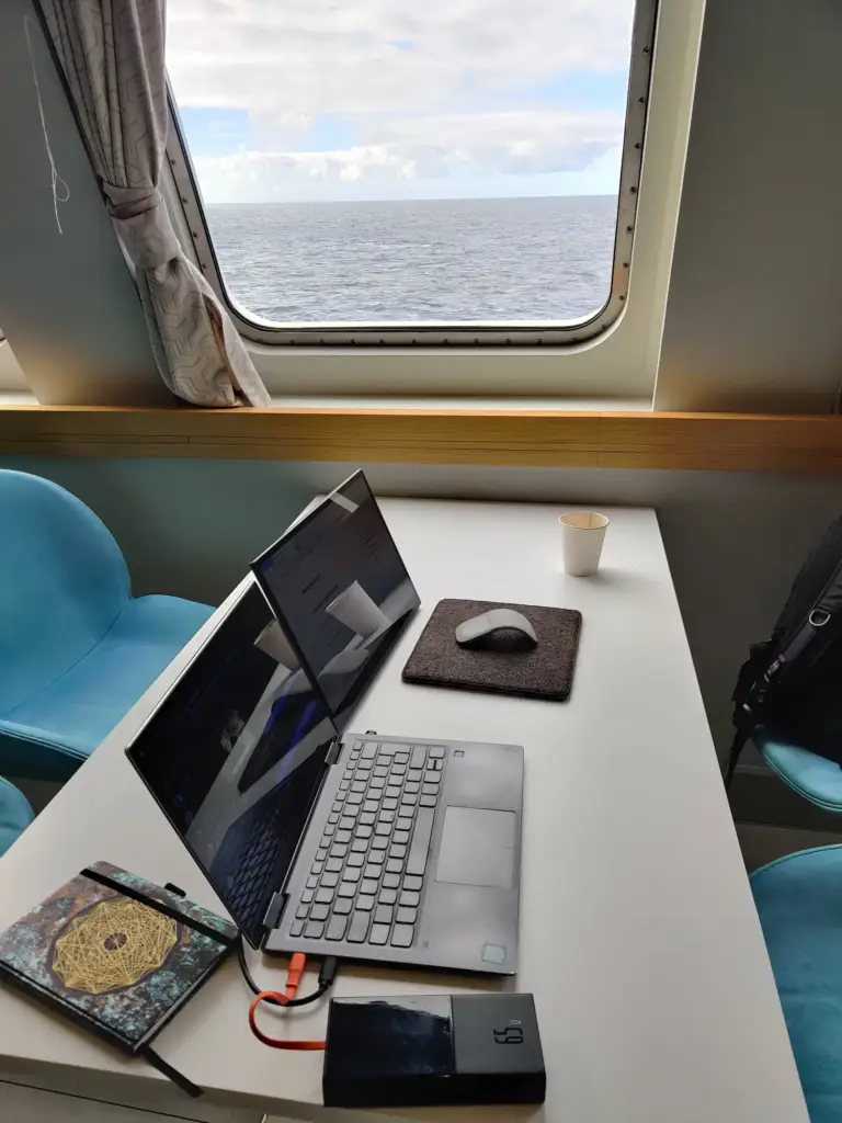 Office auf hoher See - Michi Laptop und zweiter Bildschirm auf einem Tisch, im Hintergrund sieht man durch das Fährenfenster auf das offene Meer.