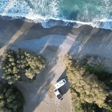 Drohenaufnahme: Zwei Wohnmobile auf einem Strand, umgeben von Pinienwäldern und im oberen Bereich sieht man die Wellen hereinrollen.